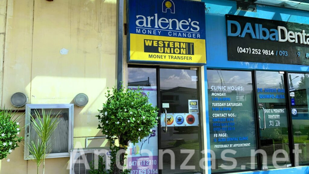 フィリピン・スービックにあるArlene's Foreign Exchange Dealer & Money Changerの店舗外観。青色を基調とした看板と下部の黄色いWESTERN UNION看板が特徴。ガラスの引き戸が入口になっており、右側には隣接する歯医者の建物も確認できる