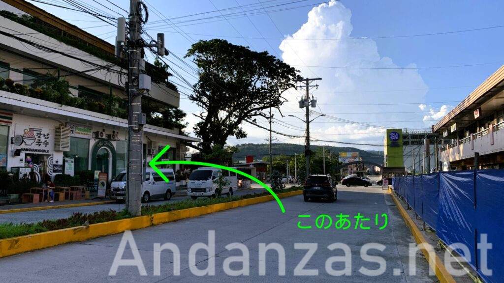 フィリピン・スービックの街並みの風景。左側には両替店舗の建物があり、緑色の矢印と「このあたり」の表示が見える。建物の影が全体に落ちる。遠くにSM City Olongapo Downtownが確認でき、青空には入道雲が浮かぶ