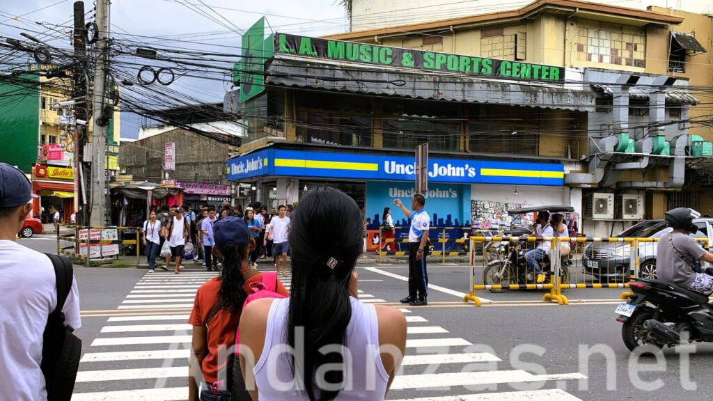 フィリピン・オロンガポで撮影した横断歩道。交通誘導員が中央に立ち、手前の女性や多くの通行人の動きを誘導。右側には青いuncles john's看板、周囲にはやや古い建物が見が並ぶ