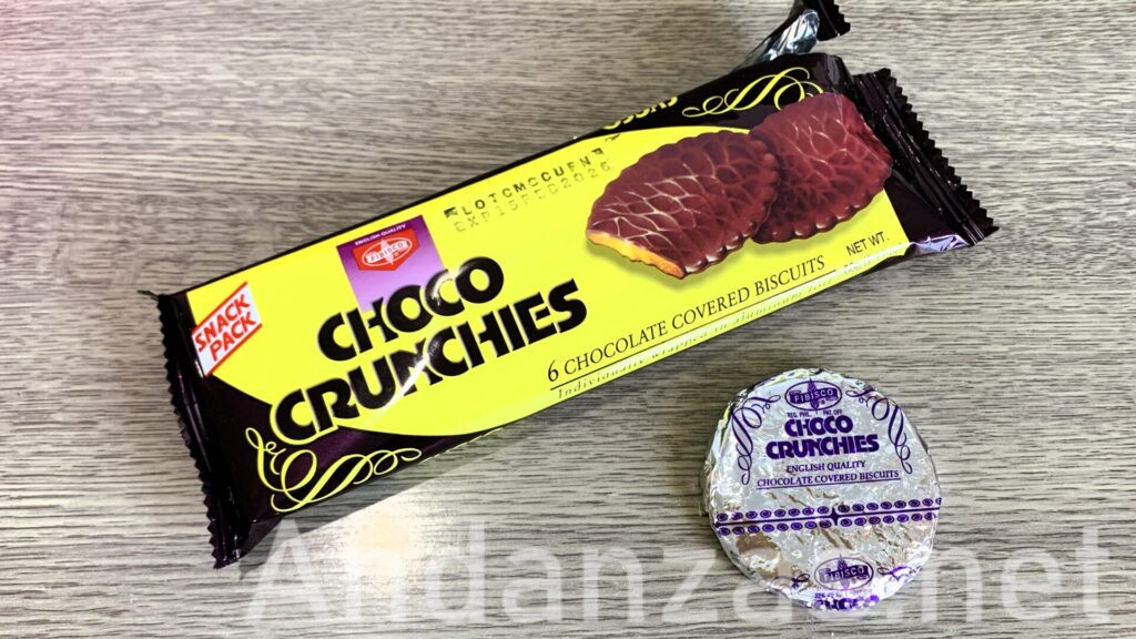 フィリピンのFibiscoブランドのChoco Crunchisパッケージと取り出された個包装のChoco Crunchis。黄色と黒を基調とした横長のパッケージが斜めに配置され、右下にアルミ製の個包装が並べられている。パッケージにはChoco Crunchisのイメージも添えられ、視覚的に特徴的なデザインが施されている