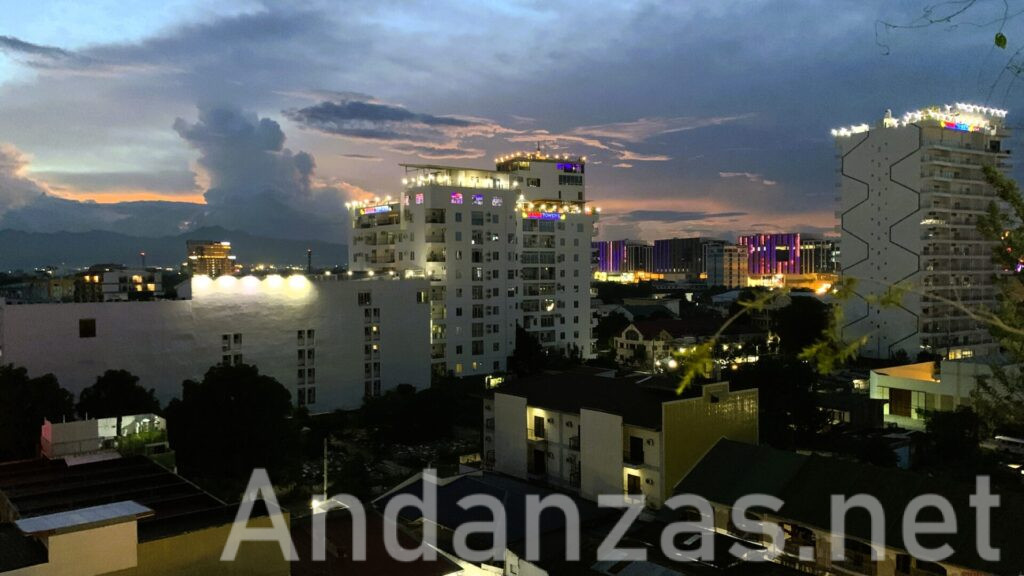 フィリピン・アンヘレスのホテル屋上から望む夕暮れの都市風景。正面と右側の建物屋上にカラフルな照明が輝き、街には低層ビルと高層ビルが立ち並ぶ。日没後の空には赤みを帯びた夕焼けとスコールを降らせた雨雲が印象的に写っている