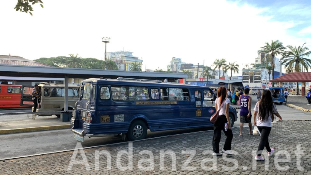 フィリピン・Bayanihan Jeepney Terminalの朝。カラフルなジプニーが停車し、奥のバス停には人々が並んで待機している。手前には、髪の長い女性2人とユニフォームの若い男性が歩いている後ろ姿が見える