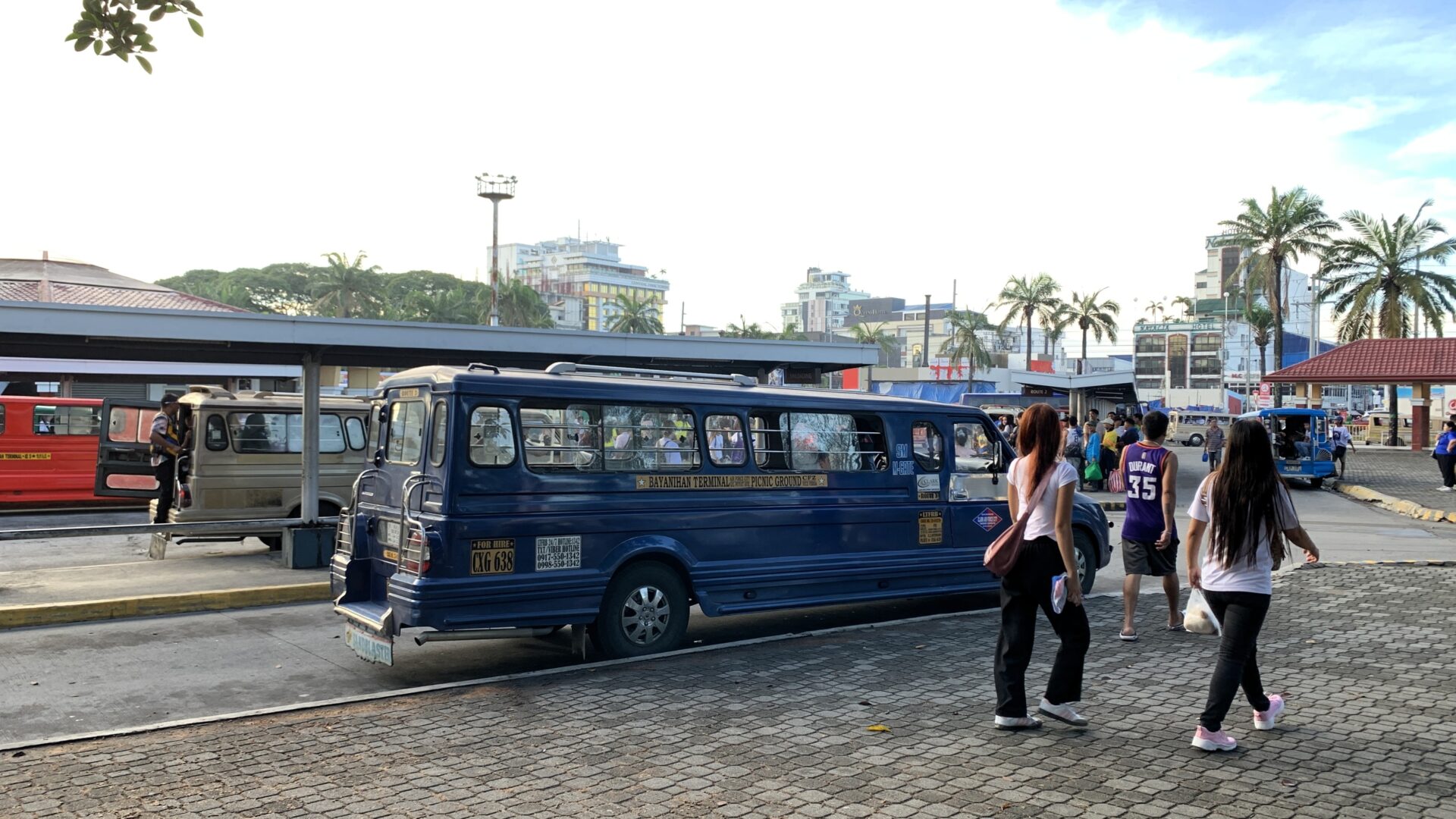 フィリピン・Bayanihan Jeepney Terminalの朝。カラフルなジプニーが停車し、奥のバス停には人々が並んで待機している。手前には、髪の長い女性2人とユニフォームの若い男性が歩いている後ろ姿が見える