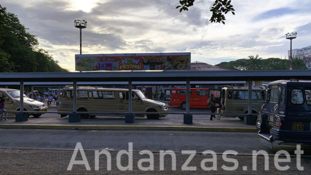 Bayanihan Jeepney Terminalの朝。何列かのバス停にジプニーが停車し、空は曇りがちながらも青空が見える。奥にはベンチに座る乗客の姿が小さく見える