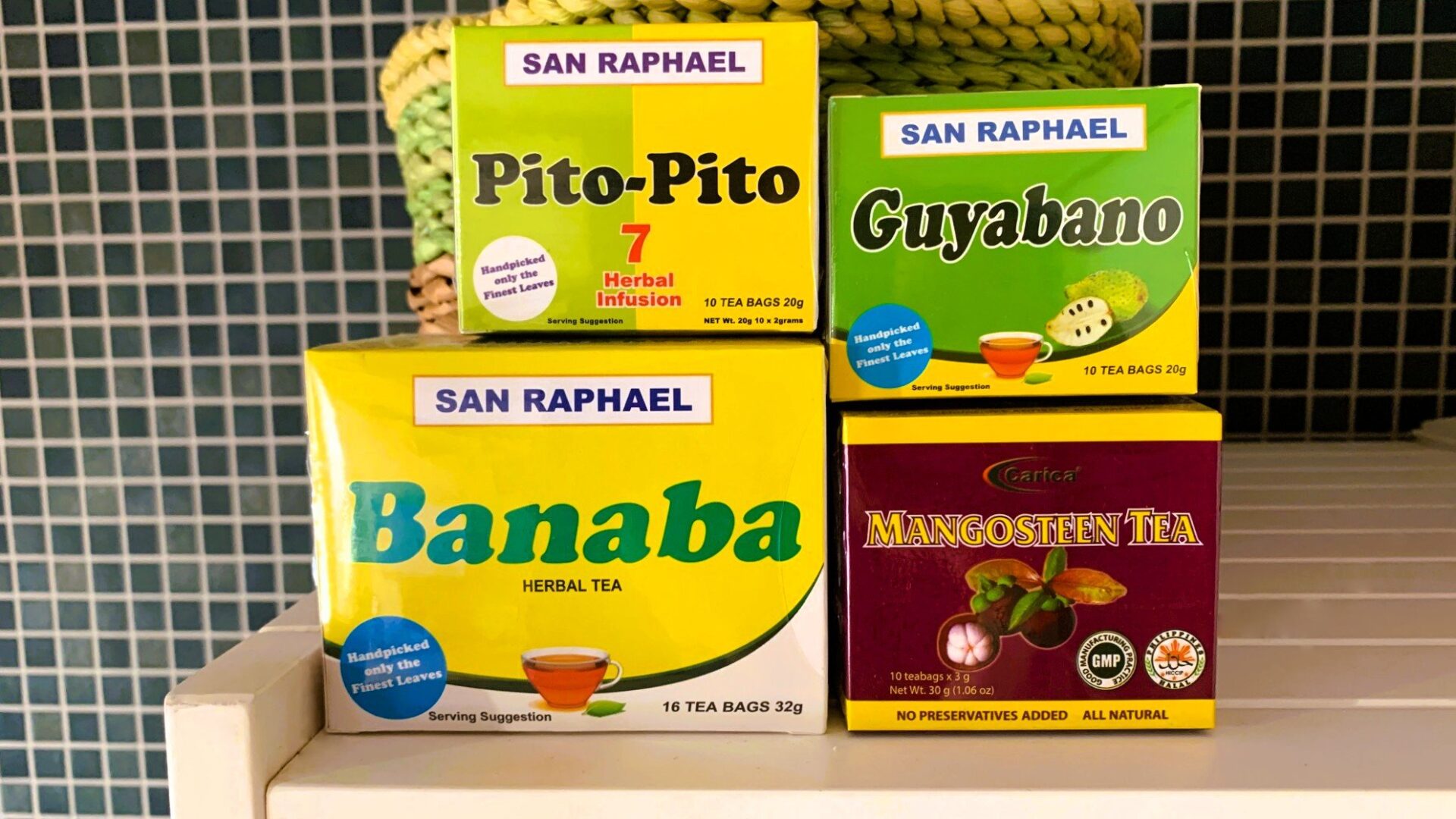 白い棚に並べられた4種類のハーブティーの箱。Guyabano、Mangosteen、Pito-Pito、Banabaの各茶があり、黄色や緑色を基調としたパッケージが多いが、Mangosteenのみ濃い赤紫色のパッケージ。背景に濃い緑色のタイルが見える