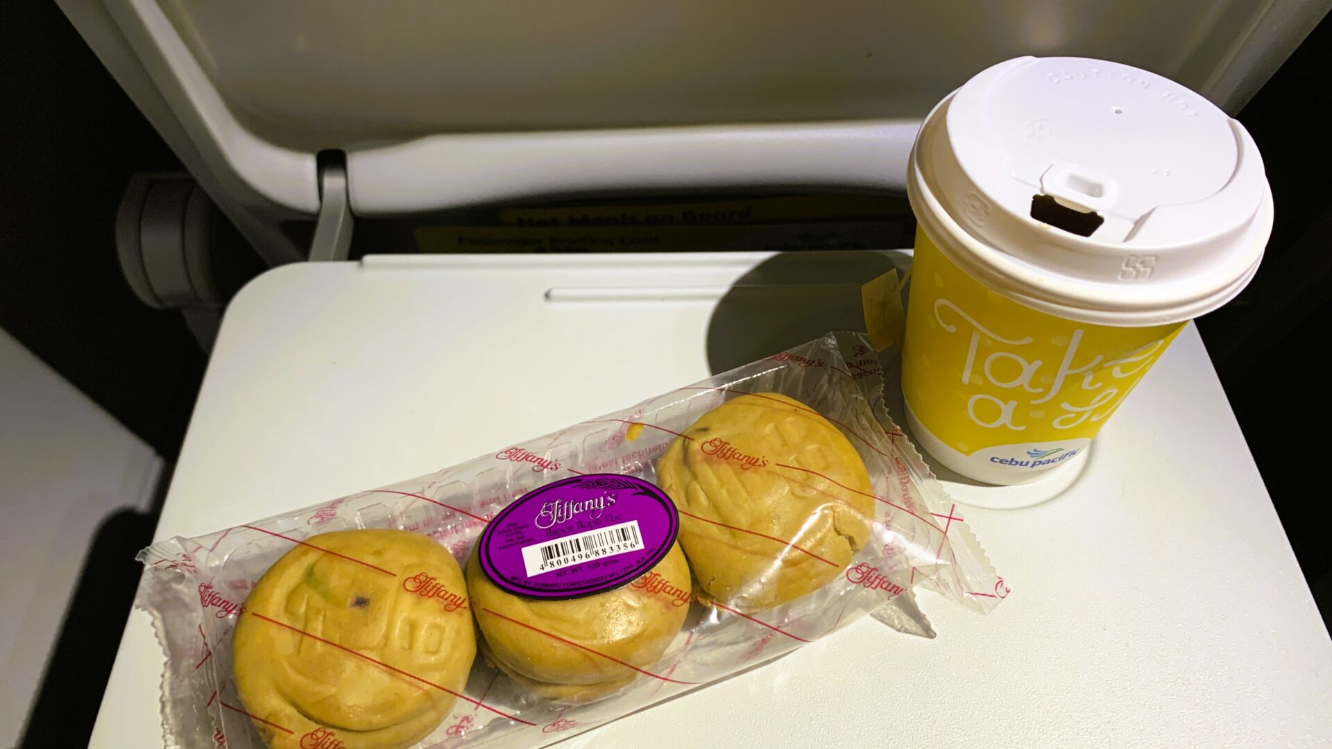 飛行機内の座席テーブルに置かれたフィリピンのHapon Hopia Ubeとコーヒー。横長の透明パックに丸い形のお菓子が3つ並び、カップ入りのコーヒーが隣に置かれている