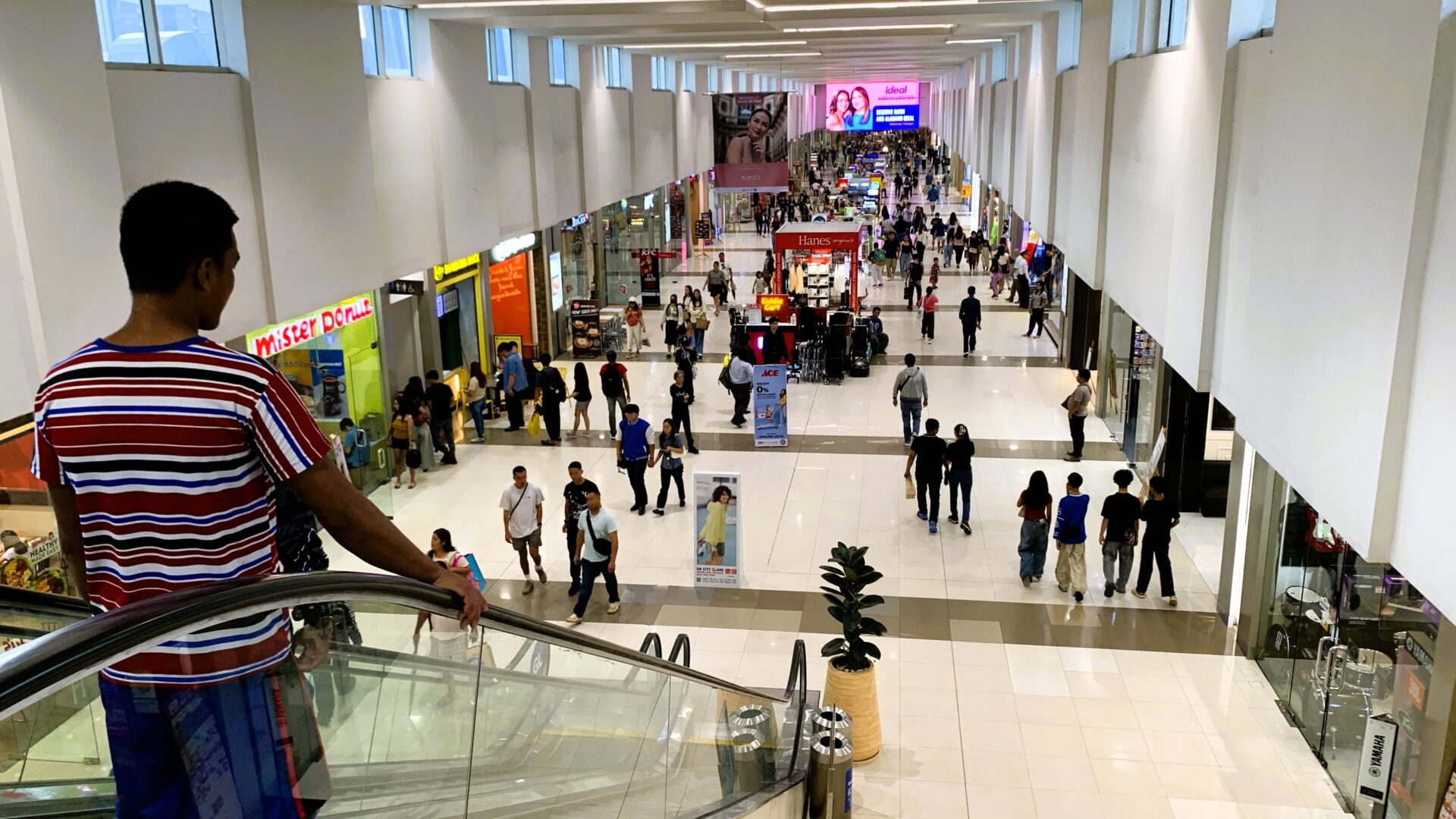 フィリピンSM City Clarkの明るい館内。白い天井と壁、両側に店舗が並び、多くの人々が歩いている。左側にはエスカレーターで下る男性が写る