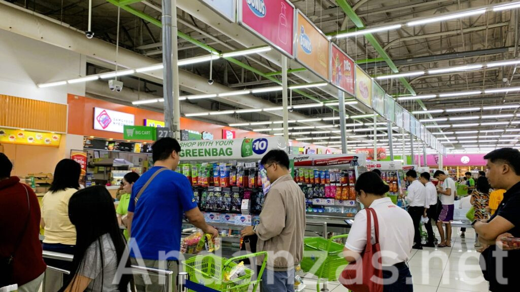 フィリピンSM Hypermarketのレジ前。たくさんの人々が順番を待っている。レジ前には商品棚が並び、蛍光灯の明るい光の下、白いタイルの床が広がっている