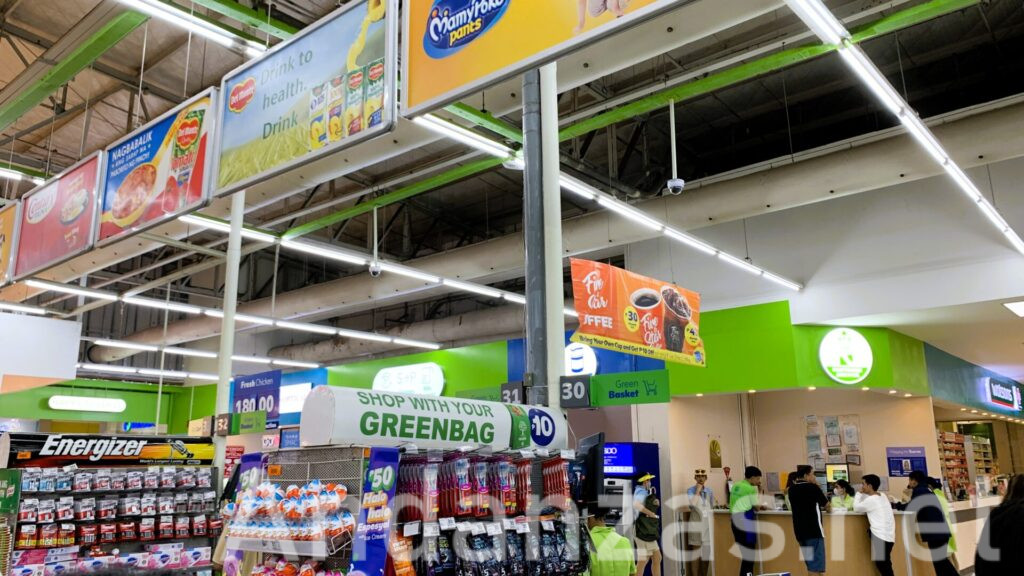 フィリピンSM Hypermarket店内風景。上部に商品の広告が並び、下部には歯ブラシやかみそり、電池やチョコレートなどが並ぶ棚が見える。奥には小店舗が並んでいる