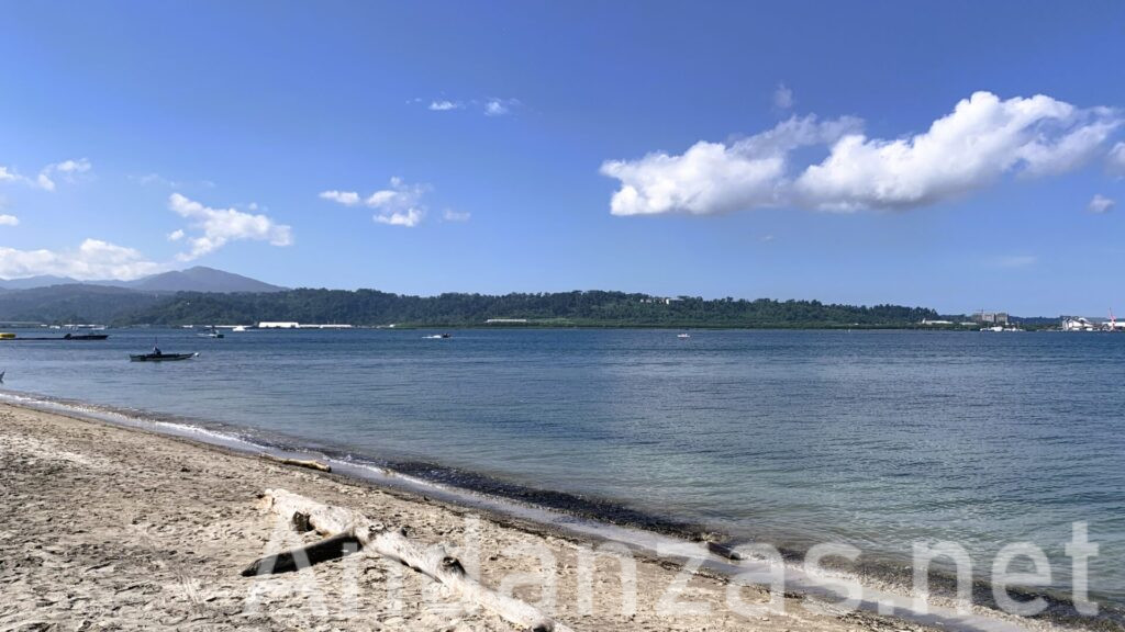 フィリピン・スービック湾の透明な海と砂浜。砂浜の手前には流木が置かれており、遠くには緑の木々と山々が広がり、青空が広がる穏やかな風景。