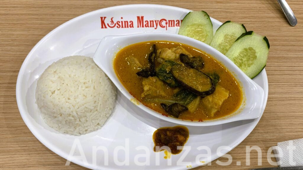 フィリピン・アンヘレスのレストランKusina Manyamanで提供される豚肉のカレカレ。右側のカレカレにはナスや野菜がたっぷり盛り付けられ、オレンジ色のスープが見える。左側にはライス、右上にはきゅうりのスライスが3枚並んでいる。木製のテーブルに置かれた一皿。