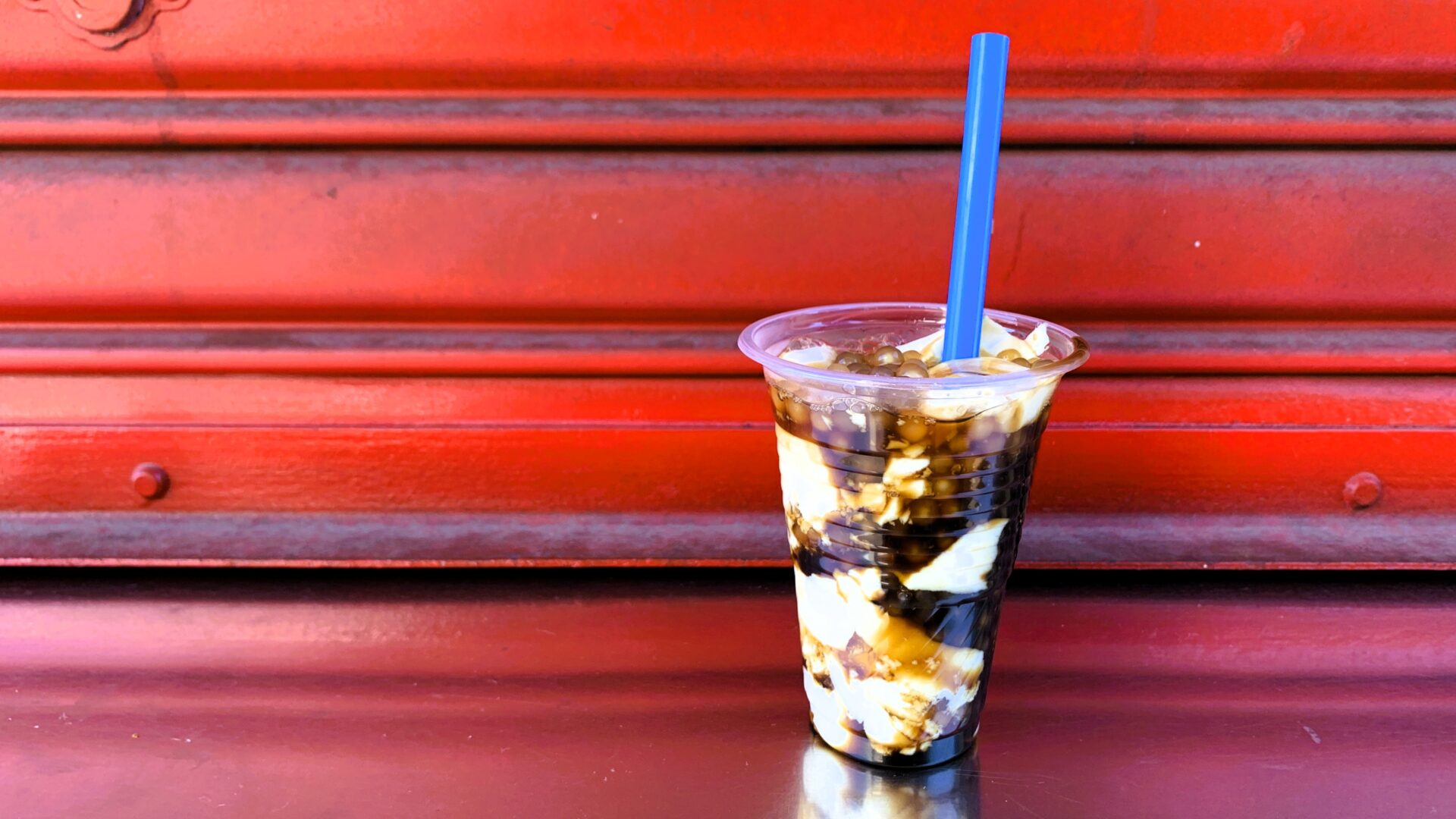 フィリピン・アンヘレスのローカル店舗でのタホ（taho）。赤いシャッターの前に、プラスチック容器に入ったソフト豆腐、タピオカ、黒蜜が横から見え、青い太めのストローが刺さっている。