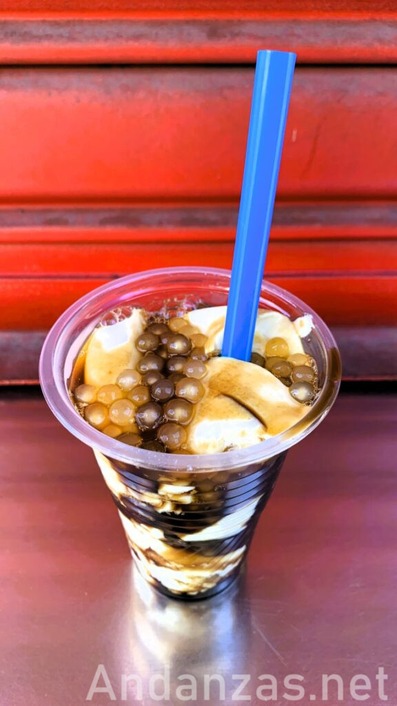 フィリピン・アンヘレスのローカルで購入したタホ(taho)。赤いシャッターの前に、プラスチック容器にソフト豆腐、タピオカ、黒蜜が見え、青い太めのストローが刺さっている。正面斜め上から撮影された写真。