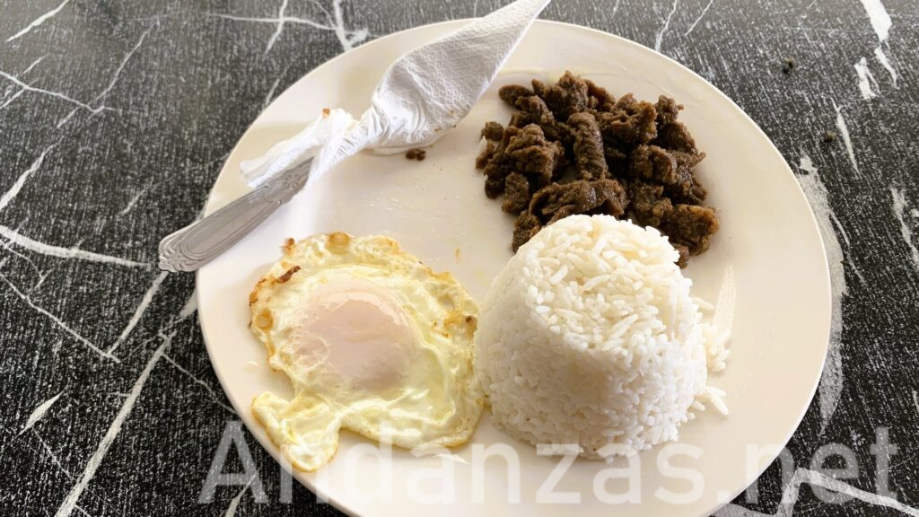 フィリピン・アンヘレスのローカル食堂で撮影したタプシログ(Tapsilog)プレート。プレートは右に大盛ライス、左に目玉焼き、右上には焼き肉が乗っている。左上にはナプキンに包まれたスプーンが添えられている。黒地に白い模様が入ったテーブルに置かれる。