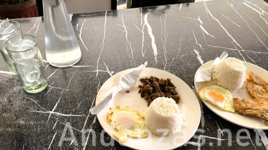 フィリピン・アンヘレスのローカル食堂で撮影した、黒地に白い模様が入ったテーブルに並べられたタプシログ(Tapsilog)とチキンのプレート。手前に2つのプレートがあり、奥には水のグラスと水入り容器が置かれている。