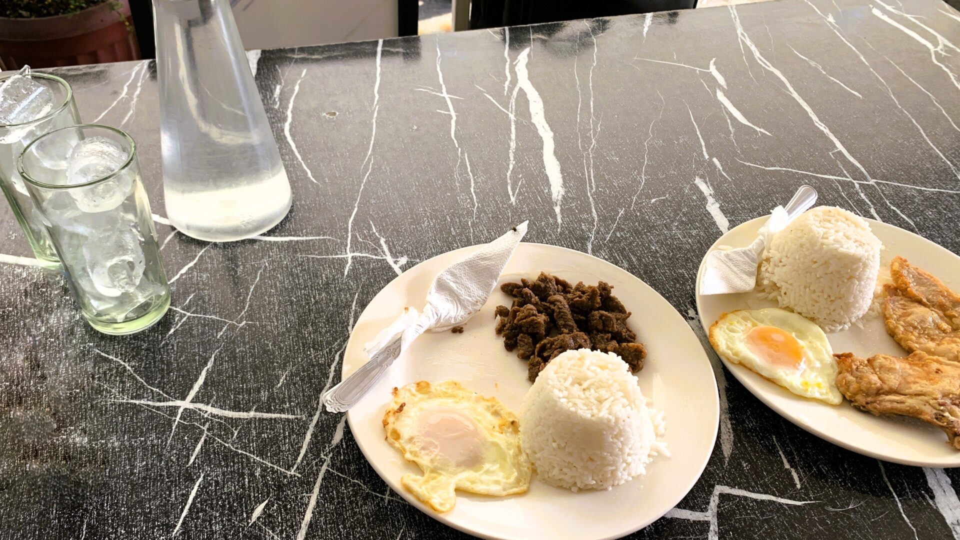 フィリピン・アンヘレスのローカル食堂で撮影した、黒地に白い模様が入ったテーブルに並べられたタプシログ（Tapsilog）とチキンのプレート。手前に2つのプレートがあり、奥には水のグラスと水入り容器が置かれている。