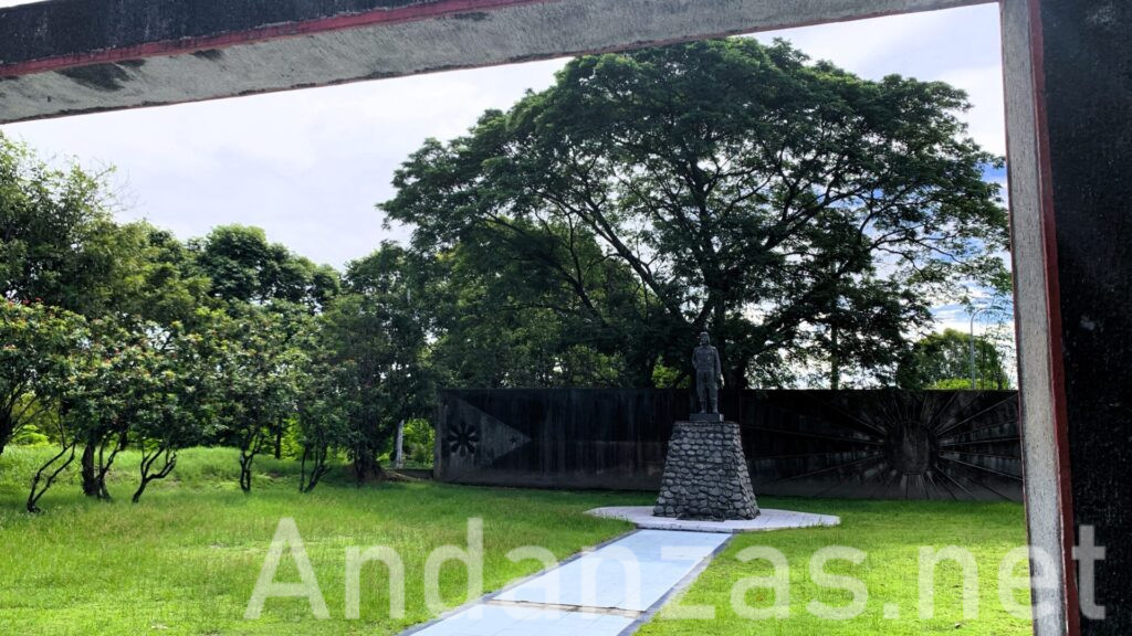 フィリピン・Kamikaze East Airfield Peace Memorialの鳥居から、斜め方向から撮影された風景。神風特攻隊の像が石の台の上に立ち、その前には白いタイルの道が長く延びる。背景には、日本とフィリピンの国旗をデザインした壁があり、周囲には緑の草や木々が広がる静かな場所。