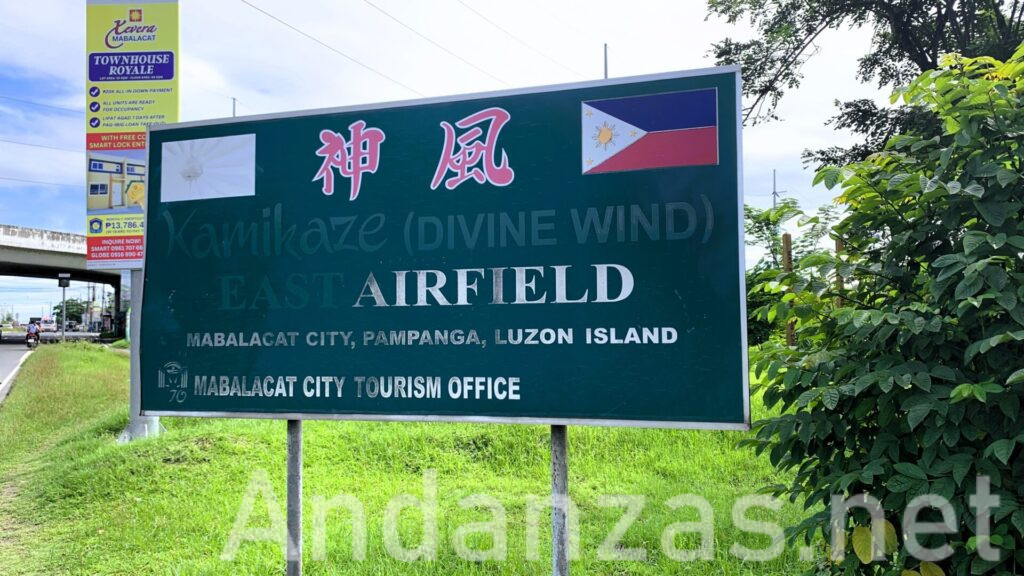 フィリピンのKamikaze East Airfield Peace Memorialにある立て看板。緑色の背景に赤字で「神風」と書かれ、日本とフィリピンの国旗が左右に描かれている。下部には英語表記もあり、周囲には植物や歩道橋が見える。