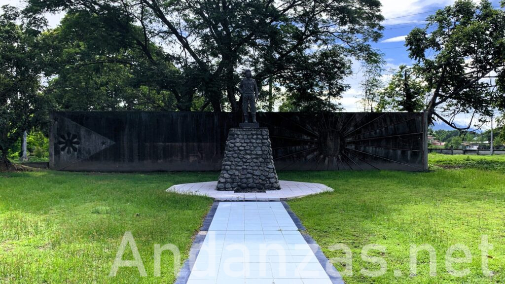 フィリピン・Kamikaze East Airfield Peace Memorialの正面からの景観。神風特攻隊の像が石の台に立ち、長い白いタイルの道が続いている。背景には日本とフィリピンの国旗が描かれた壁と緑の草、木々が見える。青空と白い雲が広がる。