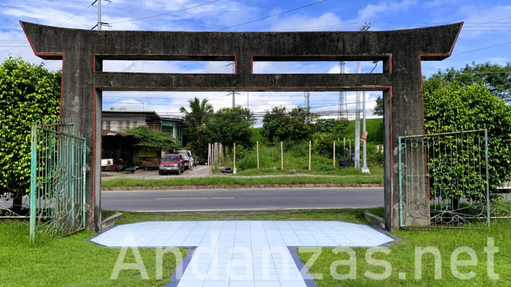 フィリピン・Kamikaze East Airfield Peace Memorialから外を写した風景。手前に鳥居があり、白いタイルの道が鳥居まで続く。奥には道路や家屋、空き地が広がり、周囲には緑の草と木々が立っている。