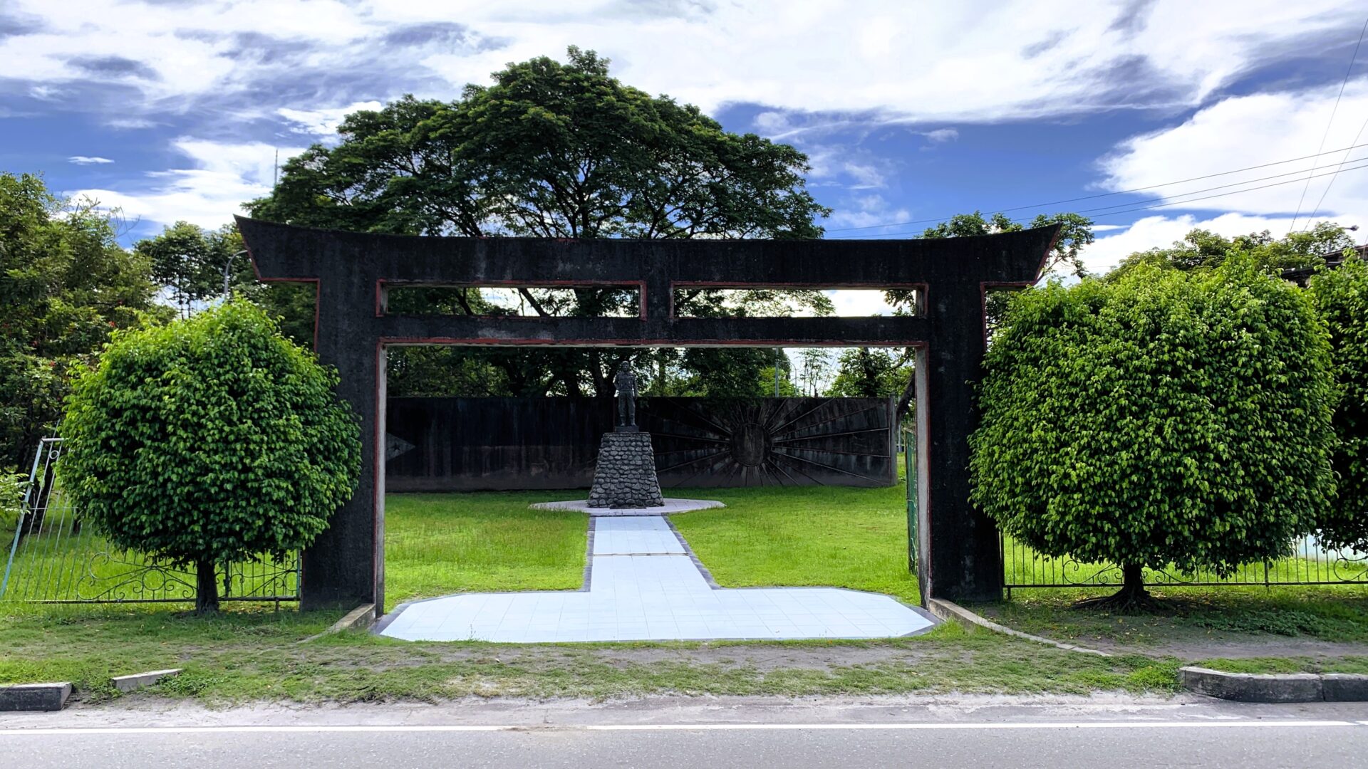 フィリピン・Kamikaze East Airfield Peace Memorialの正面から撮影。鳥居が手前にあり、奥に神風特攻隊の像が立つ。白いタイルの道が鳥居まで続き、背景には国旗デザインの壁と木々が広がる。