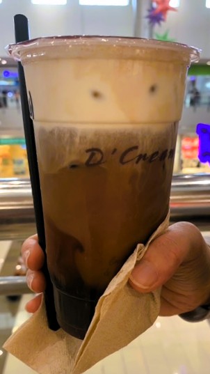 フィリピン・D'Cream Coffee & TeaのSeasalt & Cream Americano。プラスチック容器に、下部に茶色のアメリカンコーヒー、上部に白いシーソルトクリームが盛られている。ストローが添えられ、手に持たれた状態で撮影された一杯。