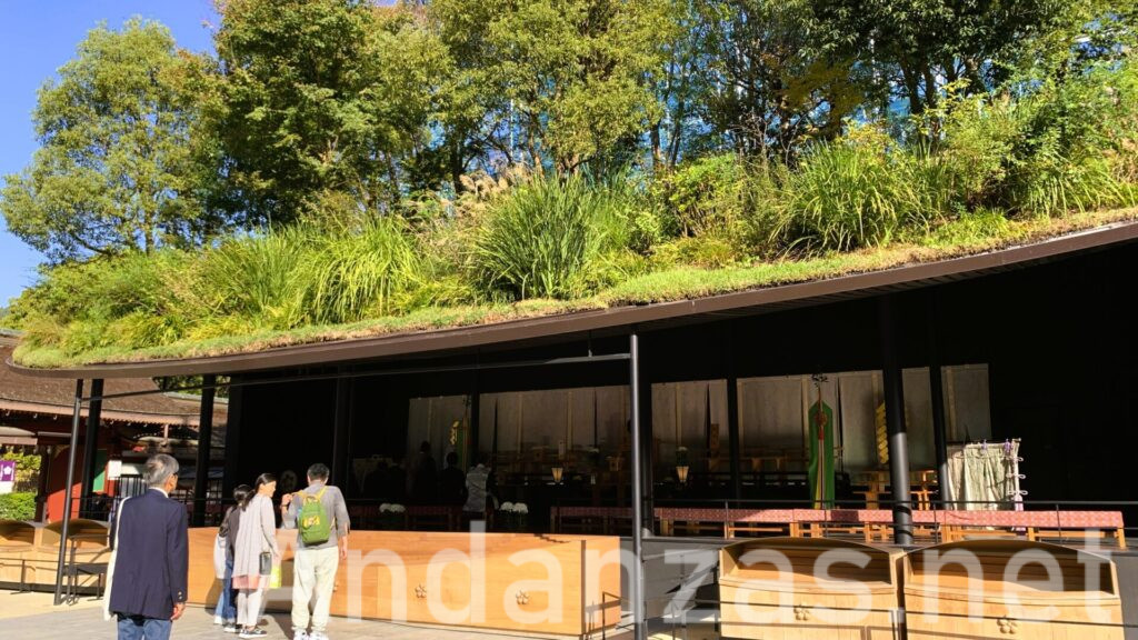 福岡・太宰府天満宮の仮殿。楕円形の屋根に木々が立つ斬新なデザインが特徴。手前には木製の賽銭箱が置かれ、晴れた空の下で緑が鮮やかに映える早朝の景色。