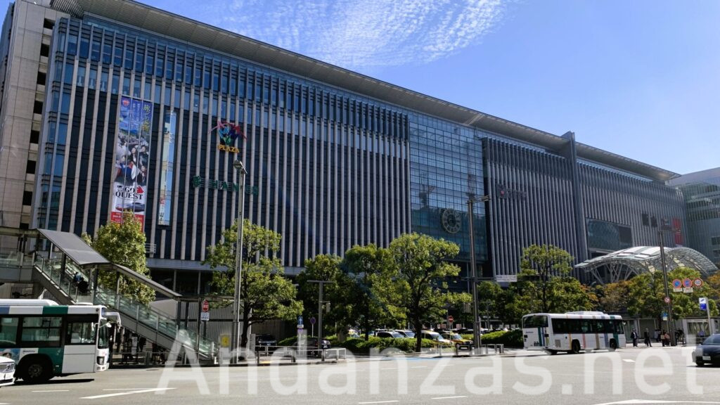 福岡・博多駅の駅ビル。正面の壁に掲げられた時計が印象的で、手前の道路にはバスや車が少し通っている。快晴の青空が広がる都会的な風景。