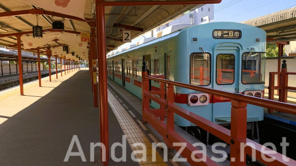 西鉄大宰府駅で撮影。右側には二日市行きの電車が、左側には駅構内が写っており、青色に赤いラインが入った丸みを帯びた車体が特徴的。駅構内の赤い柱は、大宰府天満宮を連想させる。