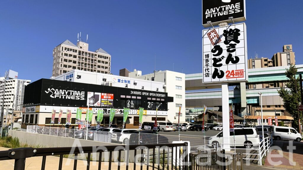資さんうどんの店舗外観。手前に「資さんうどん」の看板があり、2階建ての建物で1階が資さんうどん、2階にジムが入っている。青空が広がる快晴の日。