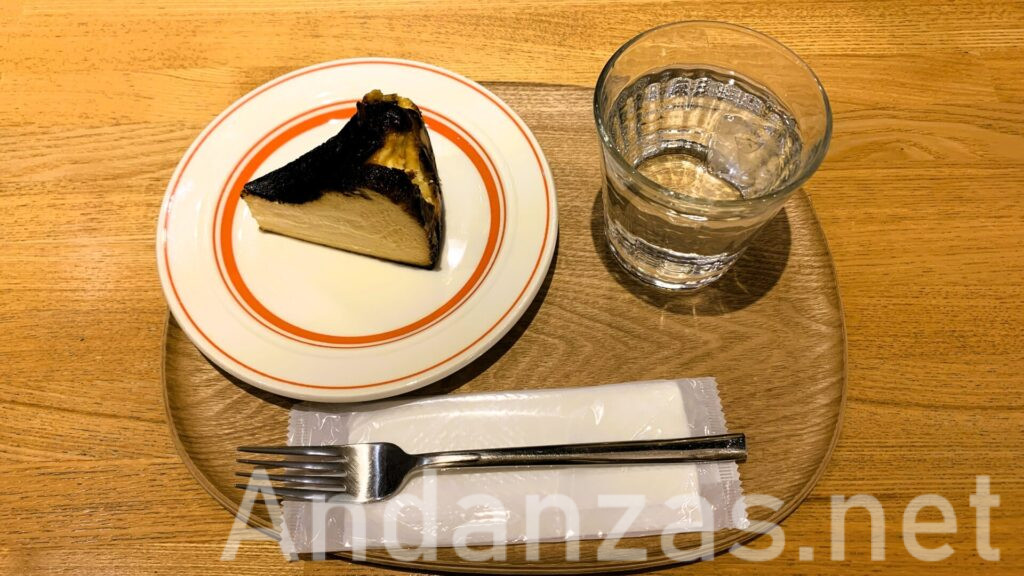 福岡・博多のATORI COFFEEのバスクケーキ。木のプレートにバスクケーキ、水のグラス、お手拭き、フォークがきれいに並べられ、木のテーブルにセットされている。バスクケーキは黒っぽい表面で、中はチーズ色。オレンジのラインが入った白い皿に盛られている。