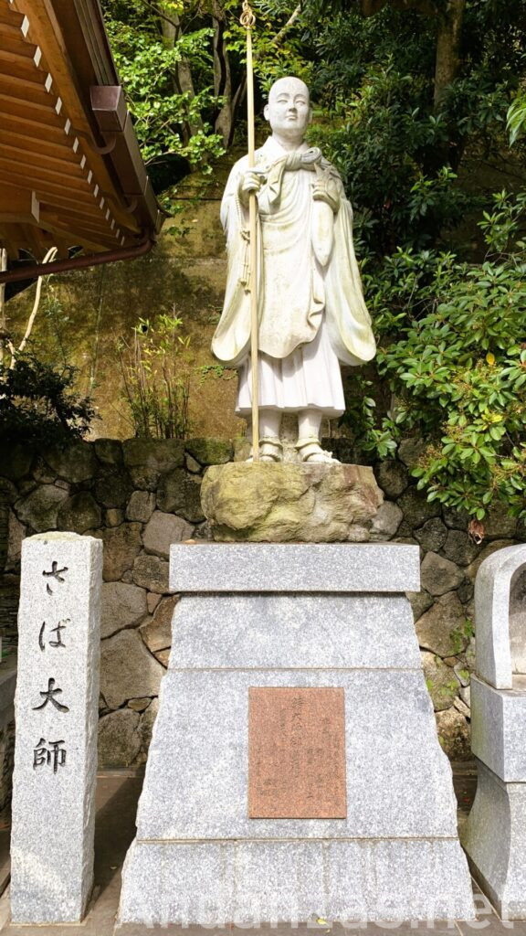 福岡・南蔵院境内のさば大師の石像。左手にさば、右手に杖を持つ白い石像。左側には「さば大師」と書かれた石碑が立っている。背景には石垣や山の崖が見える。