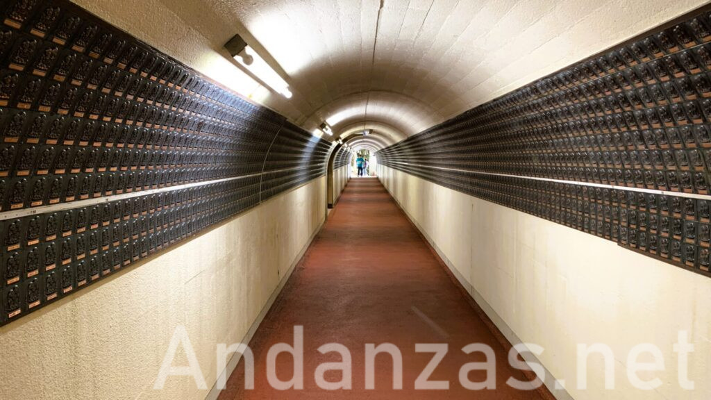 福岡・南蔵院の七福神トンネル。本堂と涅槃像を繋ぐトンネルに七福神像が祀られ、壁は白、床は朱色。先に明かりが見え、数人の参拝客が遠くに見える。