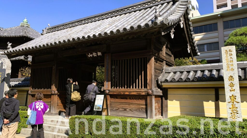 福岡・東長寺の入り口。木造の屋根瓦の建物と、右側に「東長寺」と刻まれた石が立っている。晴れた日の一枚で、入口を通過する人々が見える。