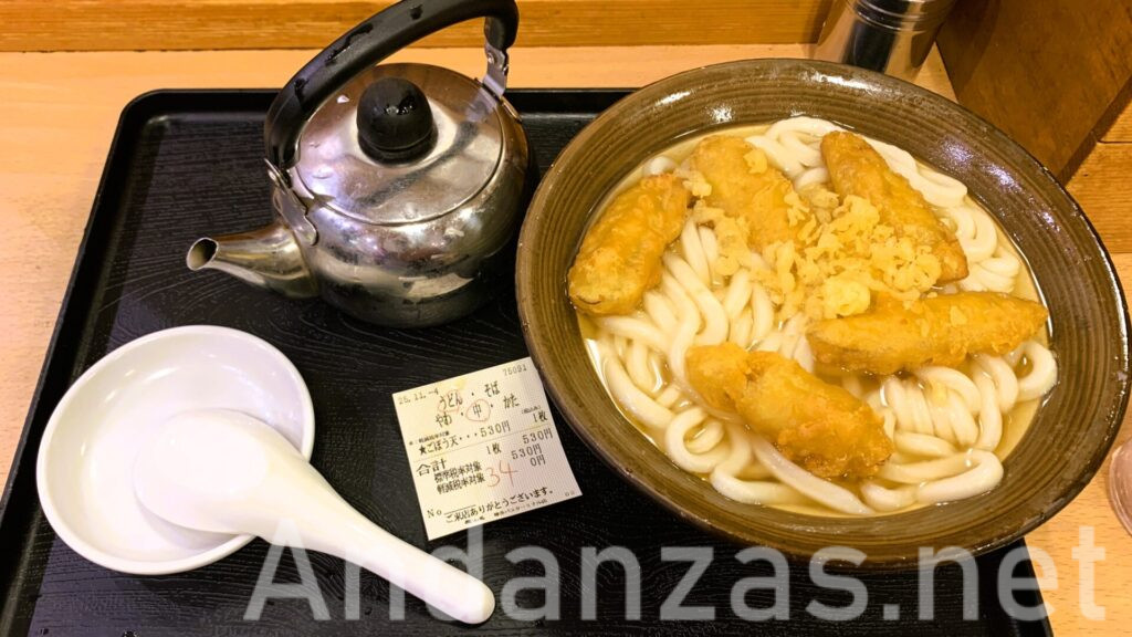 牧のうどんのごぼう天うどん。ごぼう天5つが盛られたボリュームのあるうどん。左上に小さなやかん、中央に食券、左に蓮華が置かれている。