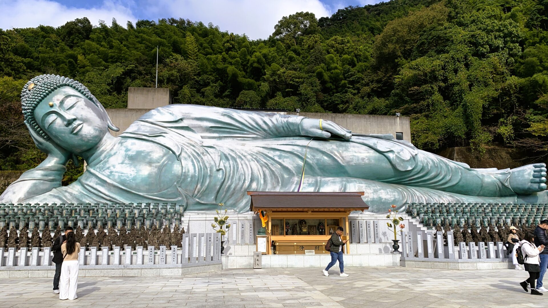 南蔵院の巨大な釈迦涅槃像。全長約41メートルの青みがかったブロンズ像が安らかな表情を浮かべ、白い石の床に観光客が何人か見える。背景には緑の木々と青空、雲も広がる。