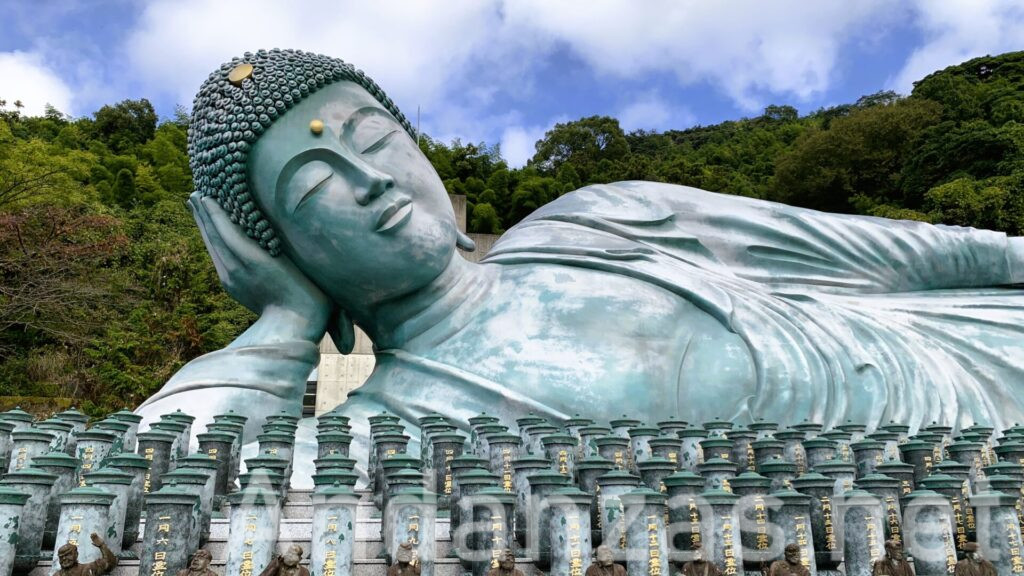 釈迦涅槃像が安らかな表情で横たわり、青みがかったブロンズ像が右手で頭を支えている。背景に山と空が見える静かな風景。