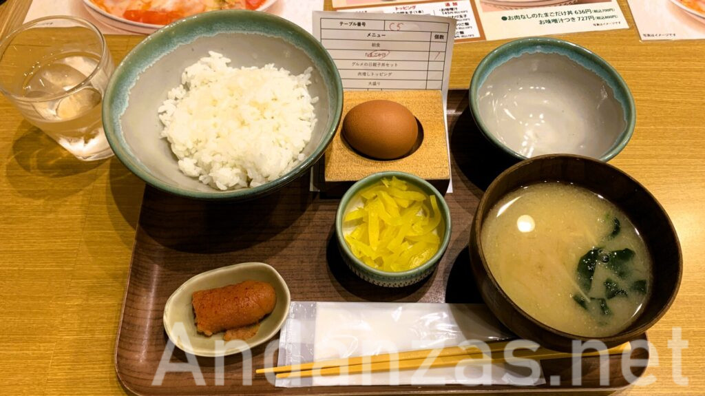 うちのたまごのたまごかけご飯。左上にご飯、右横に茶色いたまご、椀が置かれ、左下には明太子のハーフサイズ、千切りの沢庵、お手拭きが並ぶ。右にはみそ汁が添えられた木のテーブルに置かれたセット。