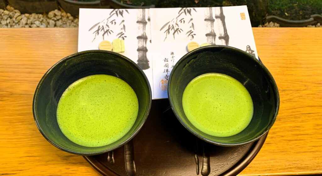 鎌倉・報国寺『休耕庵』の抹茶席。お盆の上に並べられた2つの抹茶椀と、竹の絵が描かれた和紙の上にお茶菓子が置かれ、落ち着いた茶席の風景。抹茶の緑色が美しい。