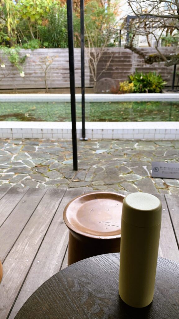 スターバックス コーヒー 鎌倉御成町店の屋外スペースを撮影した一枚。手前の木製の円形テーブルには黄色いボトルが置かれており、木製の床の奥には石の床が広がっている。さらにその先にはプールと木々が見える。テーブルの前には丸いイスが配置されている。