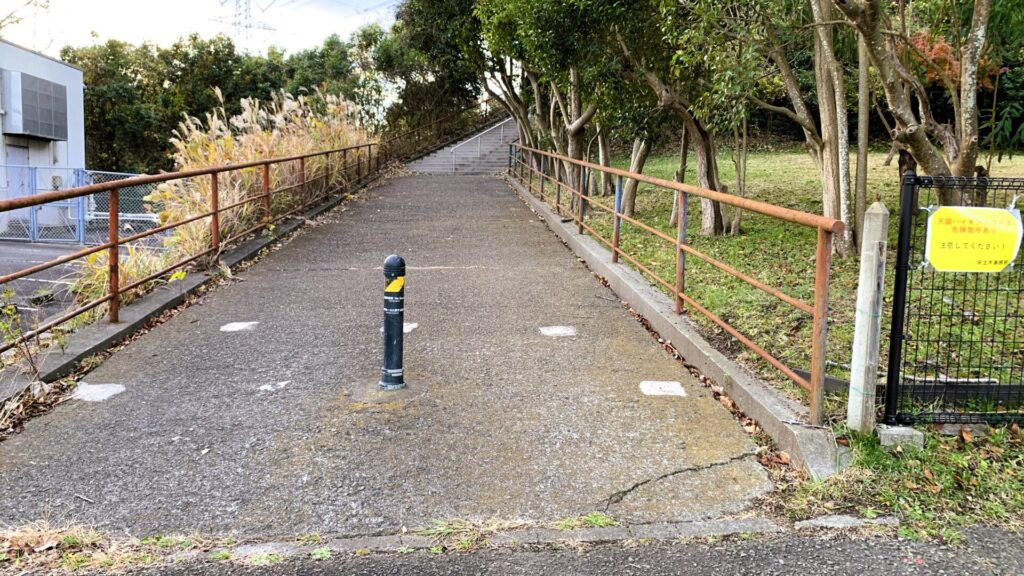 天園ハイキングコースの入口。コンクリートの道の中央には車両止めのポール、右には木々、左には建物の一部とススキが見える。奥には急な上りの階段が続いている。