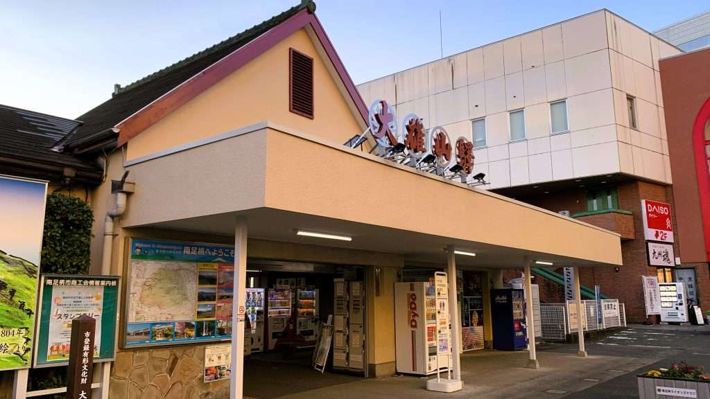 神奈川県の大雄山線の駅舎を撮影したもの。ベージュ色の外観と三角屋根が特徴的な建物で、正面に入口が見える。早朝のため人影はなく、内部には自動販売機やロッカーが設置されている静かな風景。