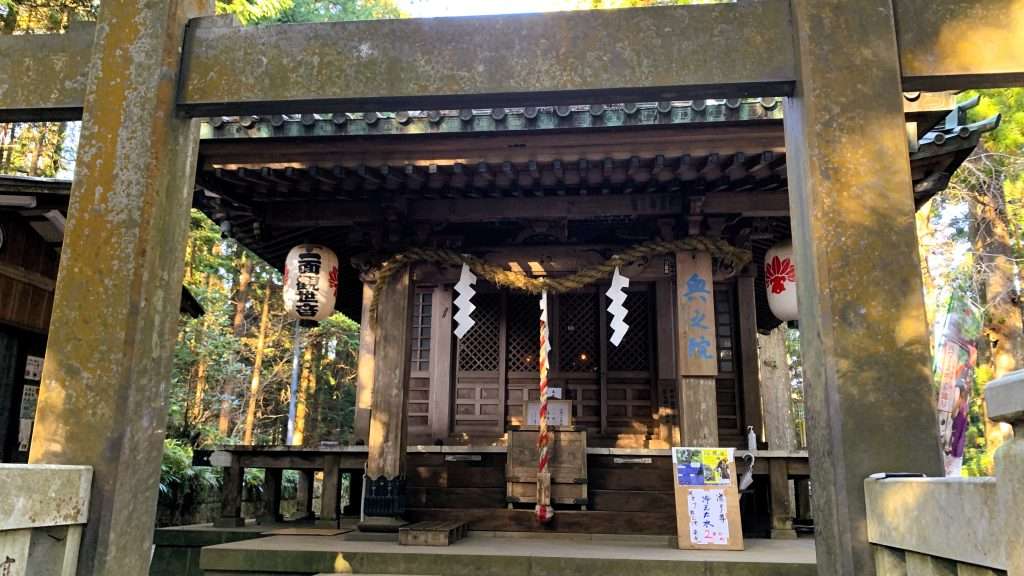 神奈川県最乗寺の奥の院の建物を撮影したものである。柱に奥の院の看板があり、中央には賽銭箱と鈴が見える。手前には石造りの鳥居が立ち、木々が建物を囲む落ち着いた景観である。