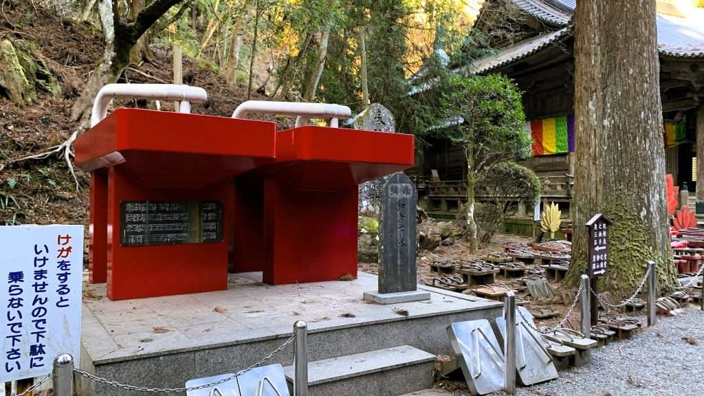 神奈川県最乗寺の高下駄が奉納されている場所を撮影したものである。左手前に巨大な赤い下駄があり、右側には小さいサイズの下駄が多数並ぶ。木製や金属製の下駄が混在し、奥には建物と山の斜面が見える。