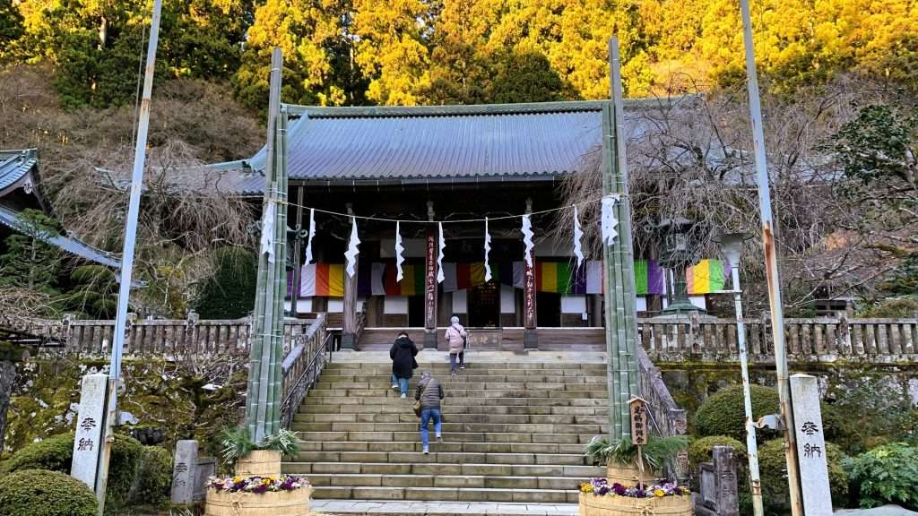 神奈川県最乗寺の本堂である。木造の建物に瓦屋根がかかり、正面には階段が設置されている。階段の手前両脇には竹の飾りが置かれ、背後には木々が広がる。三人の人が階段を上っている様子が見える。
