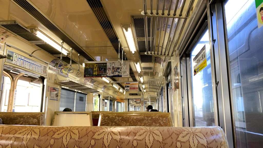 伊豆箱根鉄道大雄山線の車内を撮影したものである。手すりや荷物置き場などからレトロな雰囲気が感じられる車内で、ボックス席に座る乗客は少なめである。窓の外には黒い車体の電車が写っている。