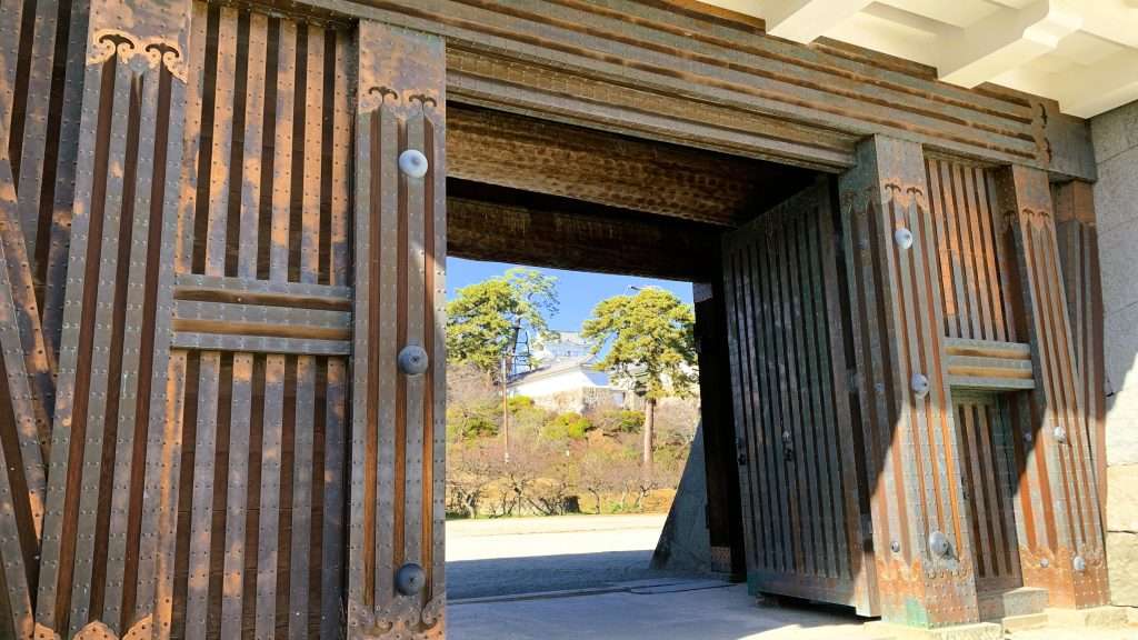 神奈川県の小田原城の銅門を下から見上げて撮影した一枚である。木製の門には銅の金具装飾が美しく配置され、門の奥には天守閣と青空、松の緑が広がる城郭の風景である。