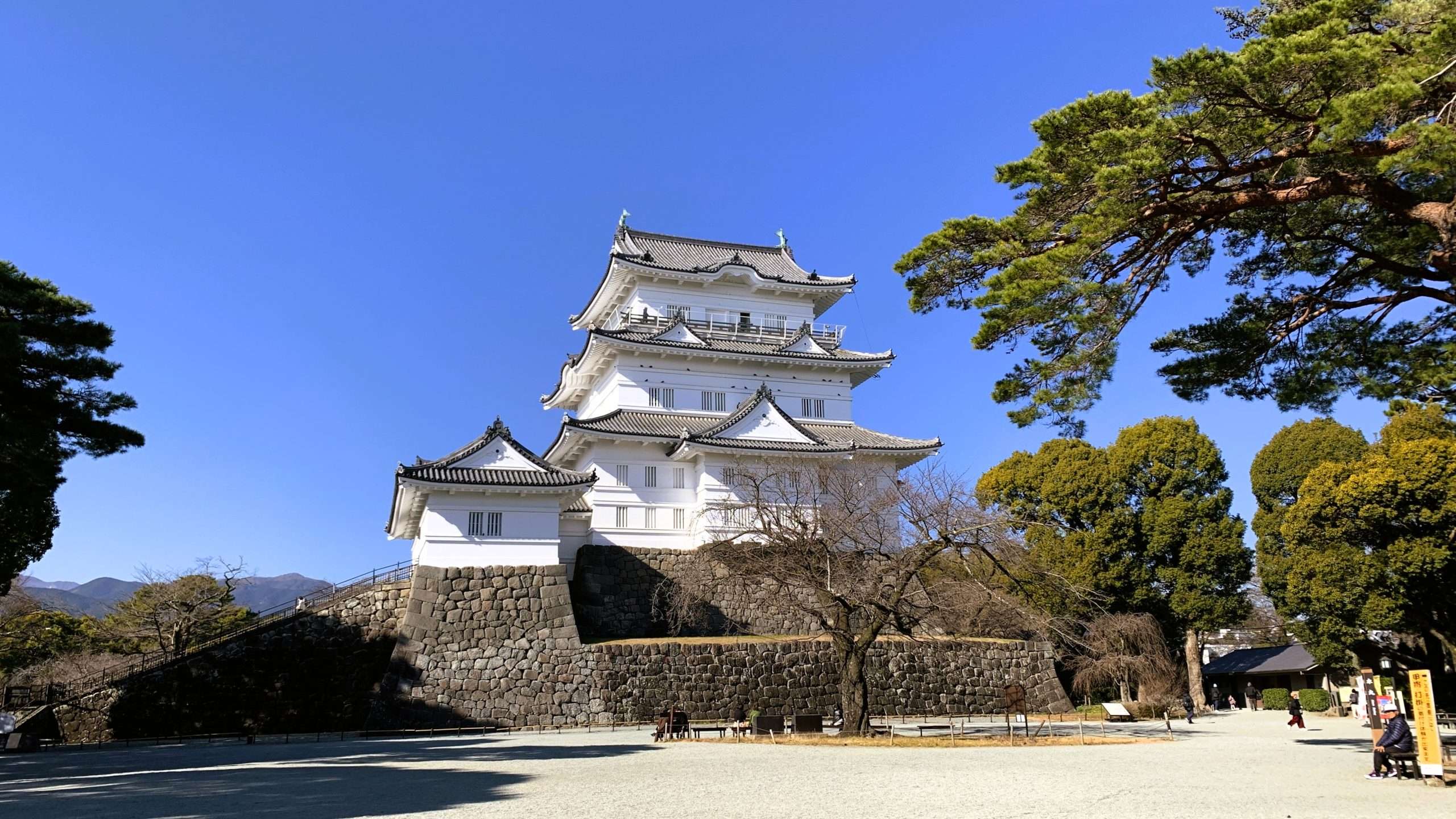 小田原城の天守閣を撮影したものである。青空の下、白い壁と灰色の瓦屋根が美しく映える。手前には広場が広がり、周囲は青々とした木々に囲まれ、人影は少ない穏やかな風景である。