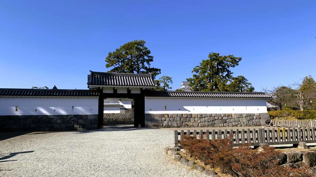神奈川県の小田原城の馬出門と周辺を撮影した風景である。青空の下に歴史ある門が立ち、右手前には柵が見える。広い敷地には砂利が敷かれ、奥には天守閣や松の木が見える静かな城内の景観である。