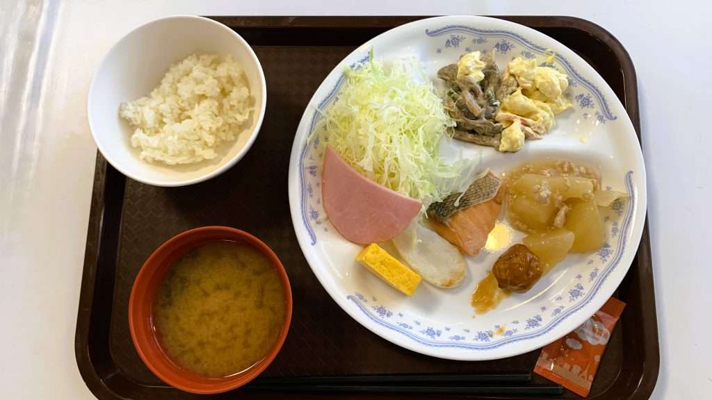 ホテルの朝食ビュッフェを上から撮影した和食スタイルの食事配置。プレートに盛られたサラダ、ハム、厚焼き玉子、焼き鮭、ミートボール、左にご飯とみそ汁が並ぶ定番の朝食バイキング。旅行や宿泊時の食事イメージ。